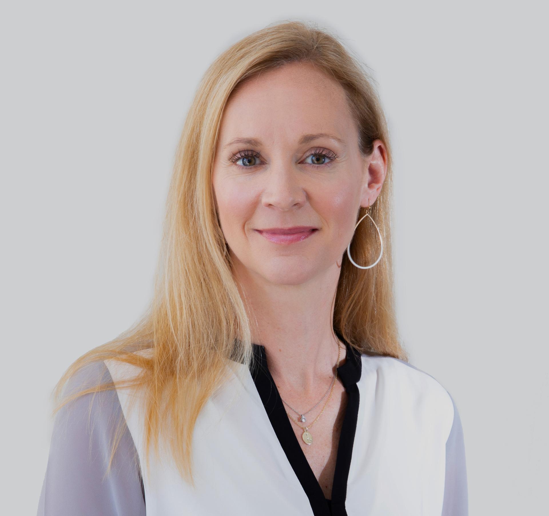A blonde white woman looking at the camera wearing a white blouse against a neutral background.